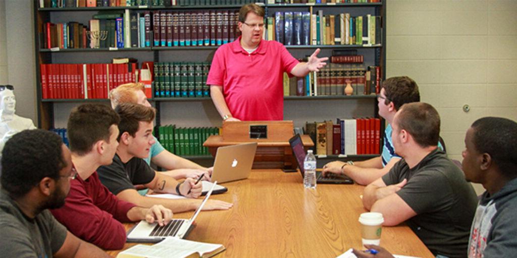 A person in a pink shirt stands at a lectern, speaking to a group of six seated people. The group is gathered around a table with books, laptops, and notebooks, in a room with shelves of books.